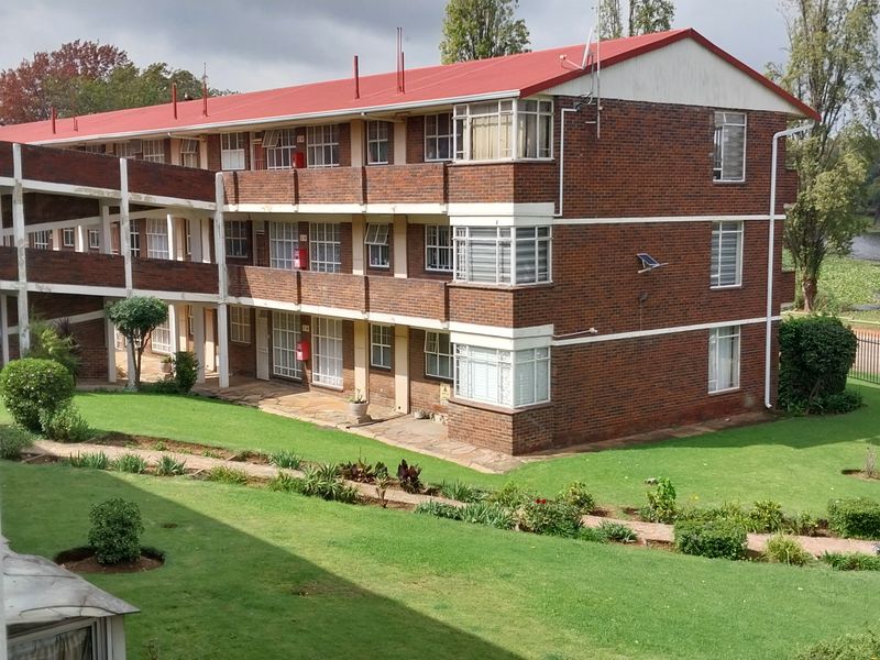 Exterior view of a multi-story brick townhouse complex featuring communal garden areas, concrete walkways, and a metal roof, situated near a water feature.