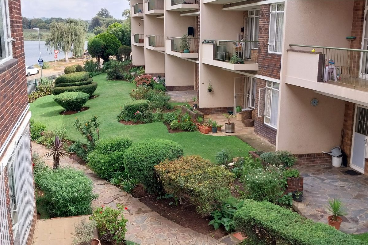Rear view of a townhouse complex featuring private balconies, paved patio areas, and a well-maintained communal garden space overlooking a nearby water feature.