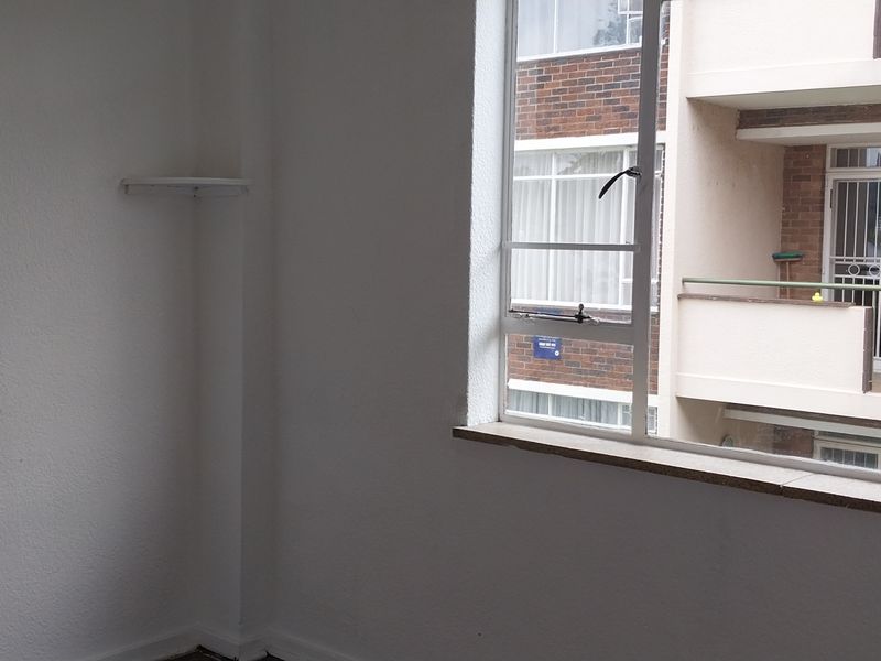 An empty bedroom featuring a large window with views of the adjacent building complex and a simple wall-mounted shelf.