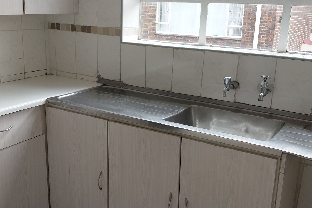 A dated kitchen area featuring laminate cabinetry, a stainless steel sink with wall-mounted taps, and a tiled backsplash, requiring cosmetic updates.