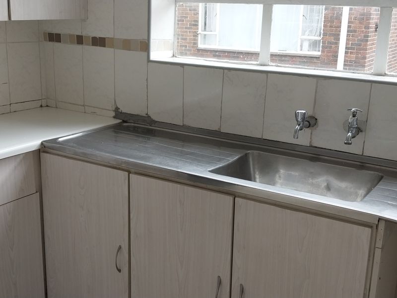 A dated kitchen area featuring laminate cabinetry, a stainless steel sink with wall-mounted taps, and a tiled backsplash, requiring cosmetic updates.