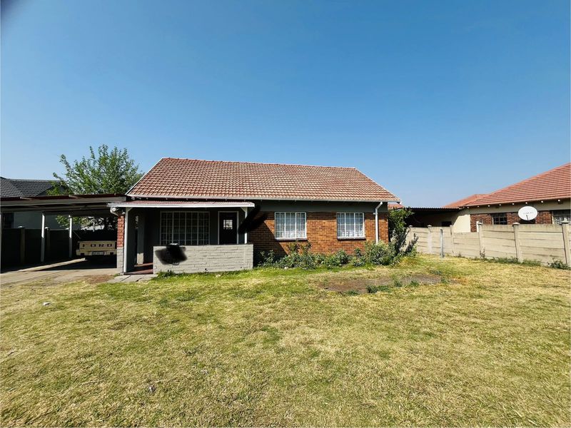 Front view of a single-story brick house featuring a tiled roof, attached carport, and a spacious front yard with boundary walling.