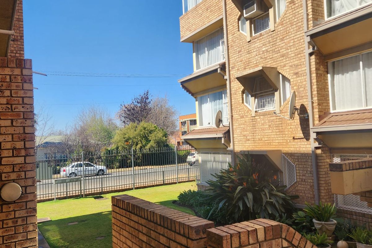 Exterior view of a multi-story apartment building featuring face brick construction, window-mounted air conditioning units, and a shared garden area with perimeter fencing.