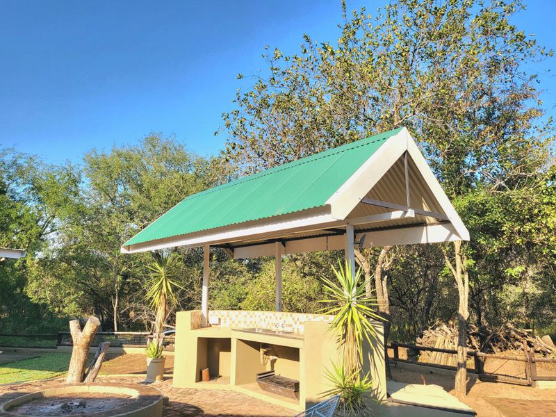 Outdoor entertainment area featuring a covered built-in braai station with a sink, tiled splashback, and brick paving, set in a natural garden environment.