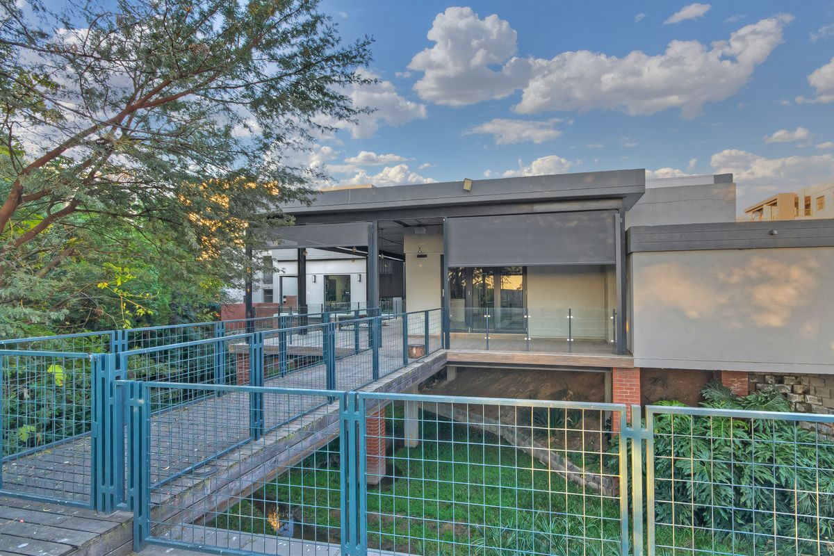 A modern elevated rear exterior featuring a wooden walkway with metal railings leading to a covered patio with glass balustrades and sliding doors.