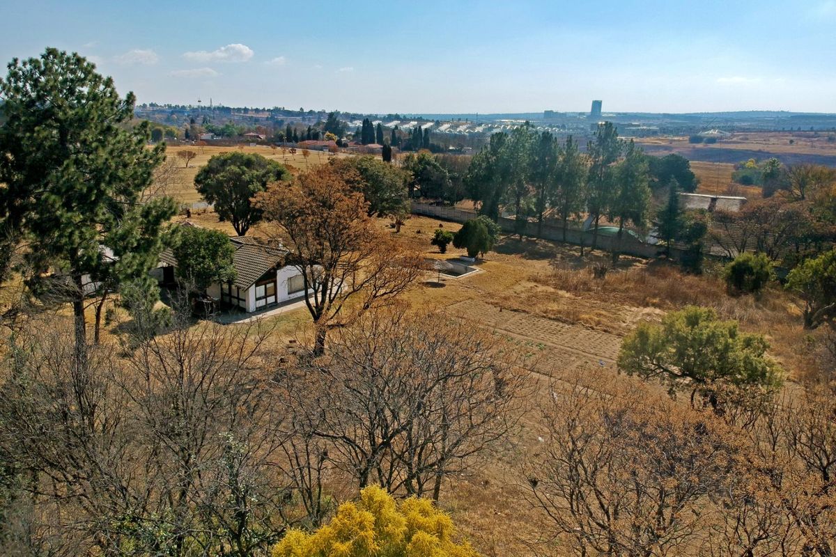 An aerial view of a large residential plot featuring mature trees, open land, and an existing dwelling with a tiled roof.