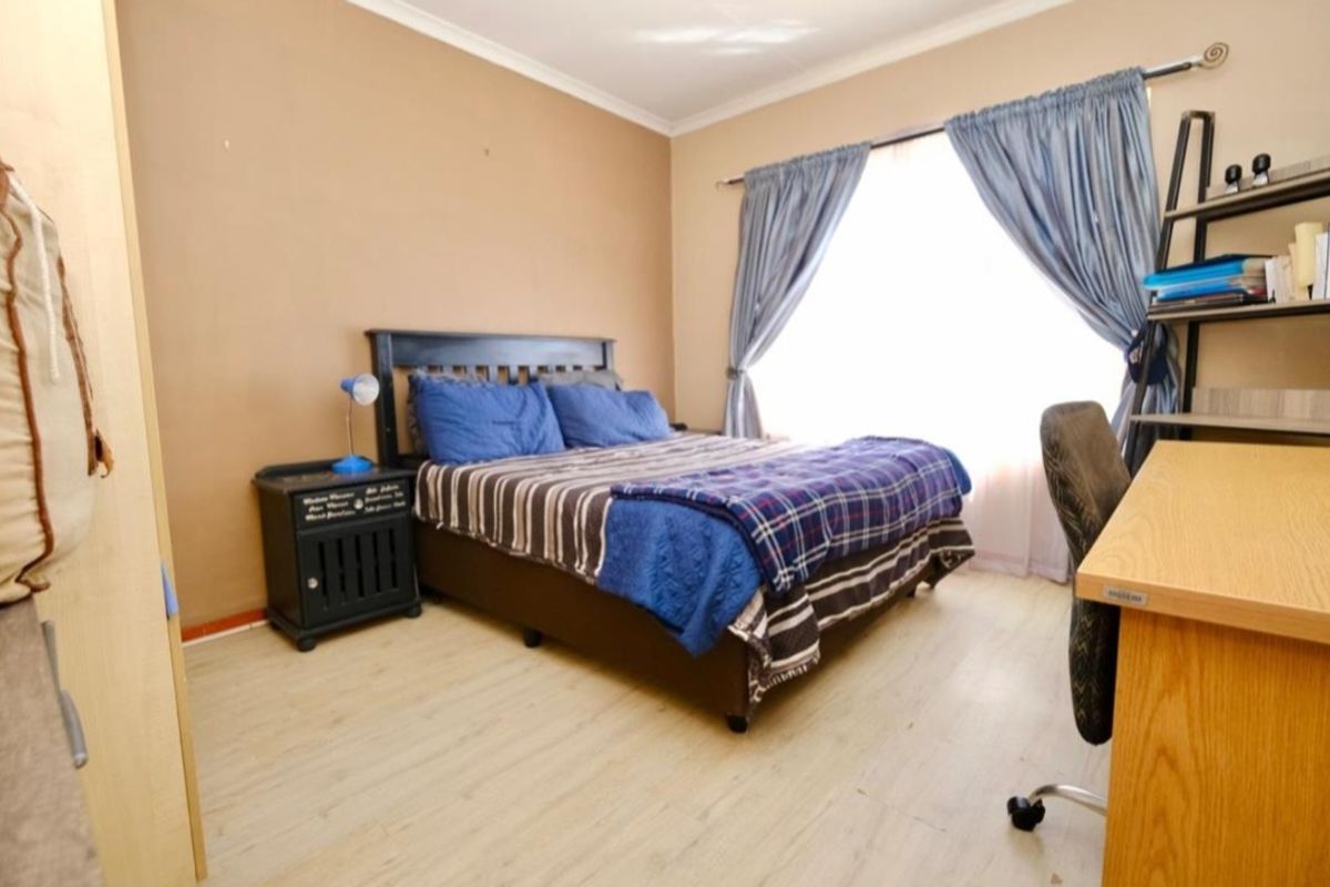 A functional bedroom featuring light-toned laminate flooring, neutral wall paint, and a window dressed with curtains.