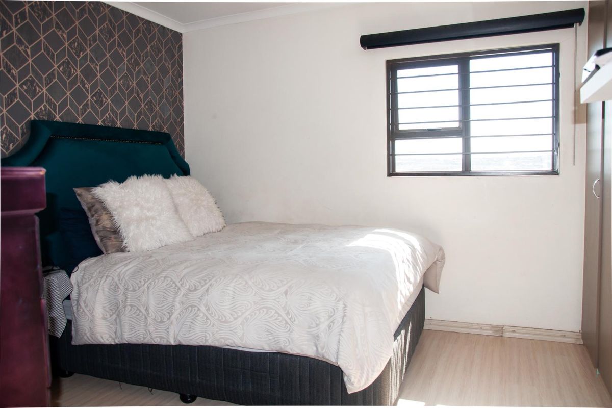 A bedroom featuring laminate flooring, a feature wall with geometric wallpaper, and a window fitted with security bars and a roller blind.