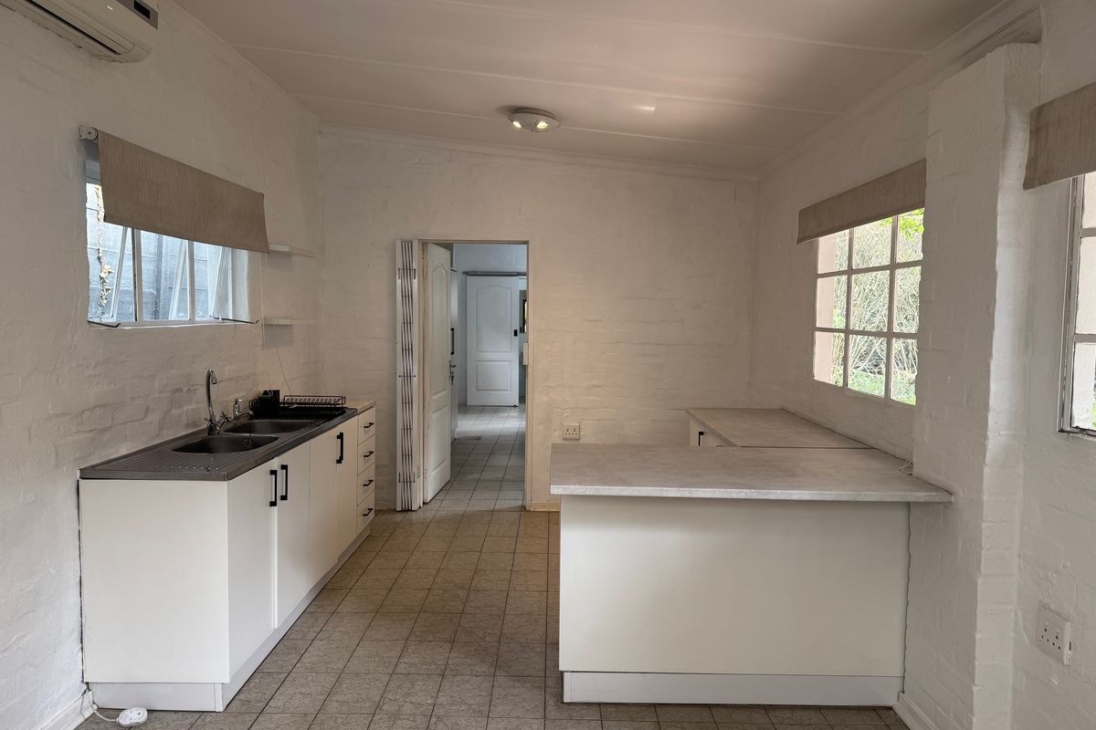 A functional, empty kitchen featuring white cabinetry, laminate countertops, a stainless steel sink, and tiled flooring, with an adjacent doorway leading to another room.