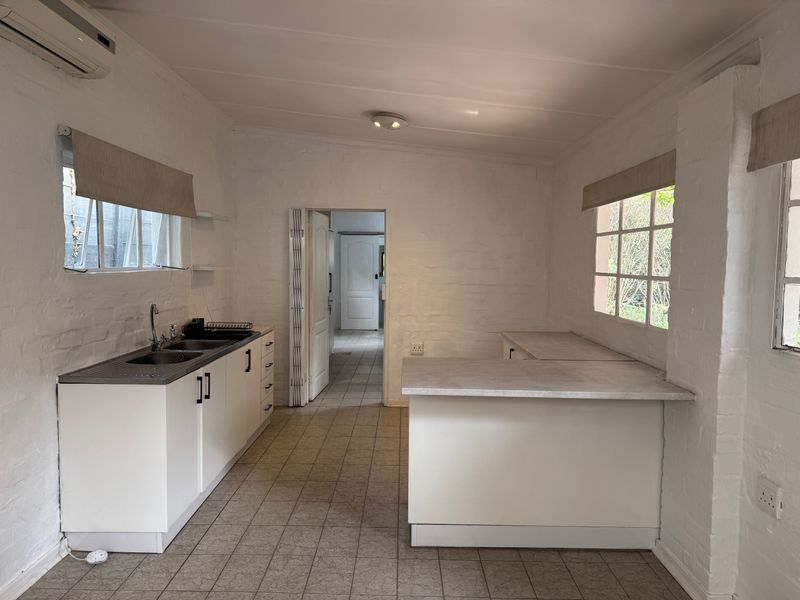 A functional, empty kitchen featuring white cabinetry, laminate countertops, a stainless steel sink, and tiled flooring, with an adjacent doorway leading to another room.