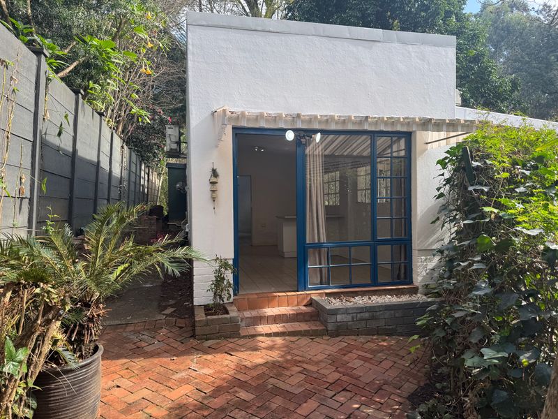 A rear exterior view of the cottage featuring a brick-paved patio, blue-framed glass sliding doors, and a small raised garden planter.