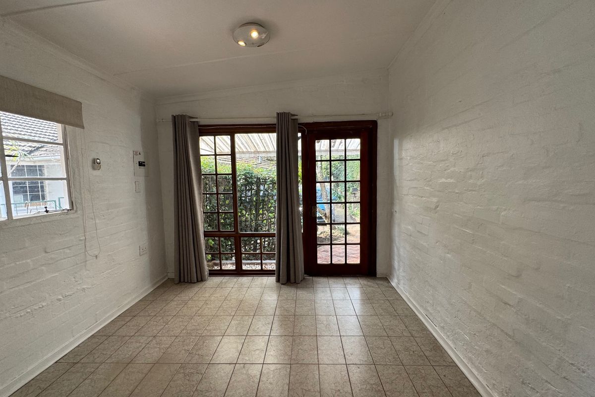 A bright, empty living room featuring tiled floors, painted brick walls, and direct access to the garden through timber-framed glass doors.