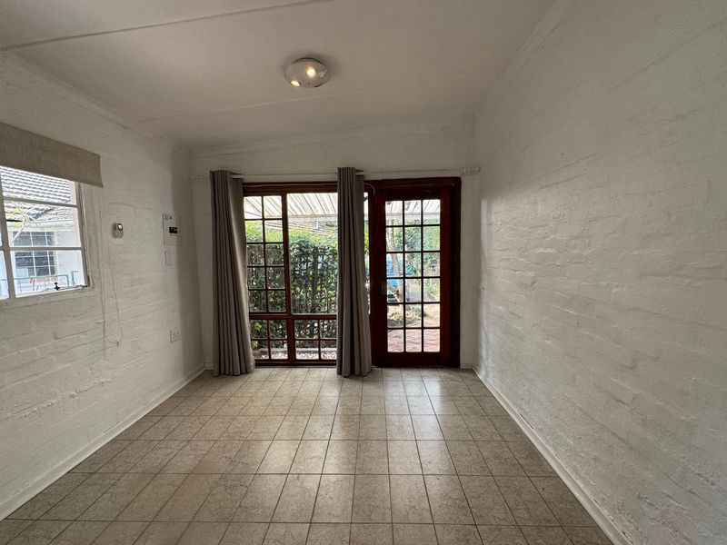A bright, empty living room featuring tiled floors, painted brick walls, and direct access to the garden through timber-framed glass doors.
