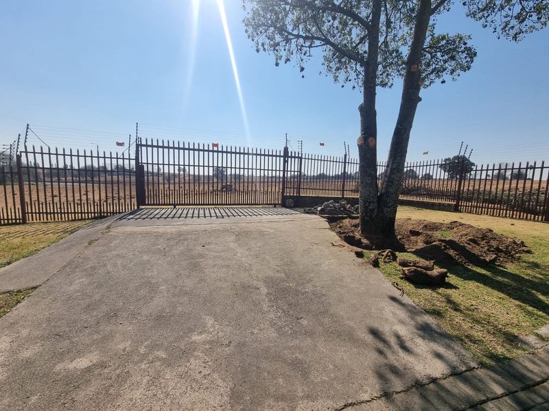 A view of the property entrance featuring a metal driveway gate, perimeter fencing with electric security wires, and a concrete driveway.
