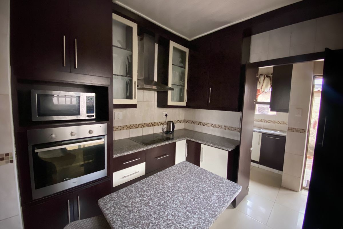 A functional kitchen featuring dark wood cabinetry, granite countertops, built-in oven, microwave, and an adjoining scullery area with tiled finishes.