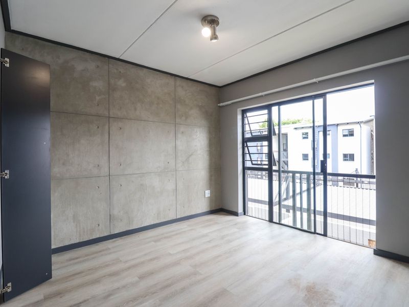 A modern, empty bedroom featuring a concrete accent wall, light laminate flooring, and a sliding glass door leading to a balcony.