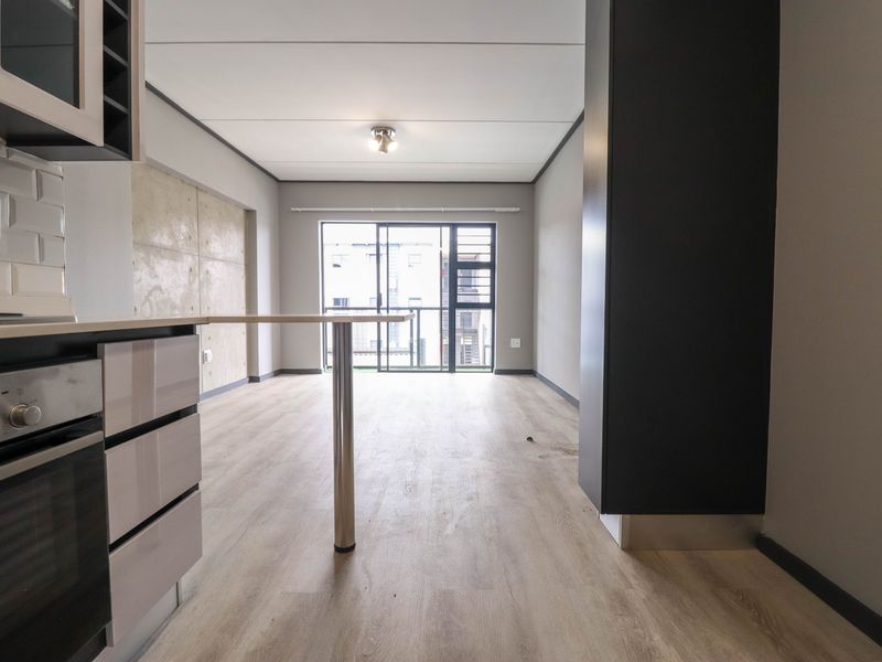 Open-plan living area featuring modern laminate flooring, a compact kitchen with built-in oven, and a sliding door leading to a balcony.