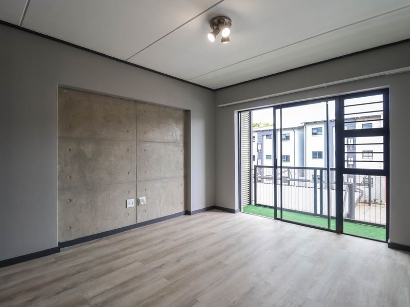 A bright, empty living room featuring modern laminate flooring, a concrete accent wall, and a sliding glass door leading to a balcony.