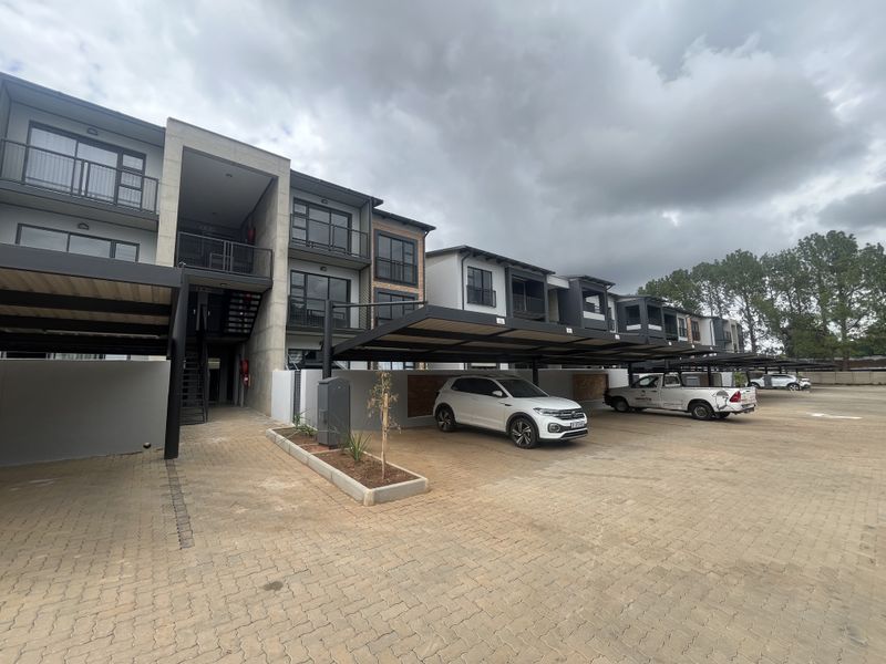 Front exterior view of a modern townhouse complex featuring paved parking areas, covered carports, and multi-level units with private balconies.