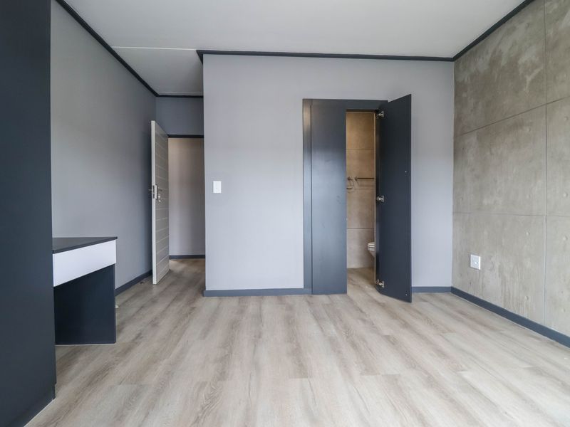 A modern, empty bedroom featuring light laminate flooring, a concrete feature wall, and a built-in desk, with an adjoining en-suite bathroom.