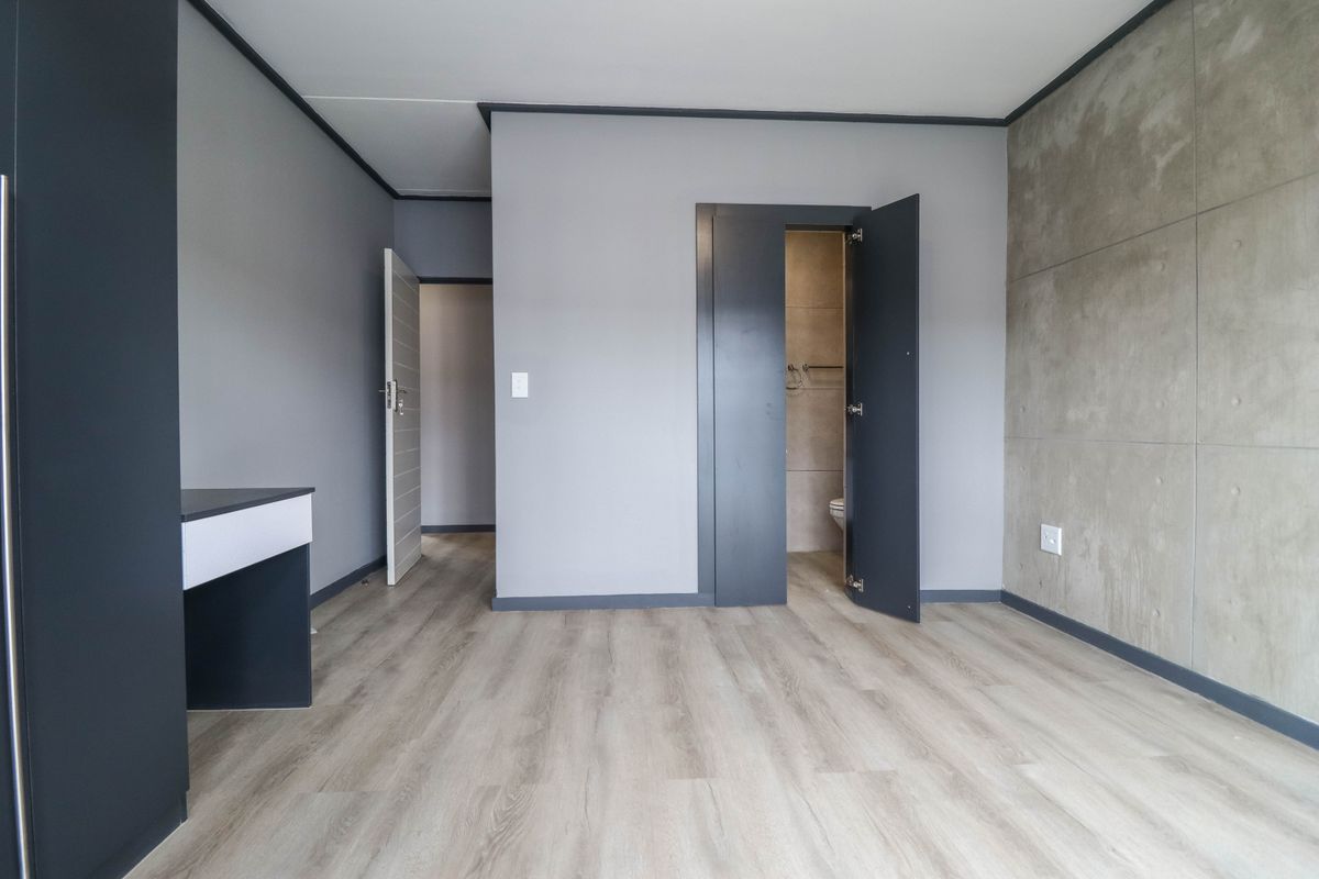 A modern, empty bedroom featuring light laminate flooring, a concrete feature wall, and a built-in desk, with an adjoining en-suite bathroom.