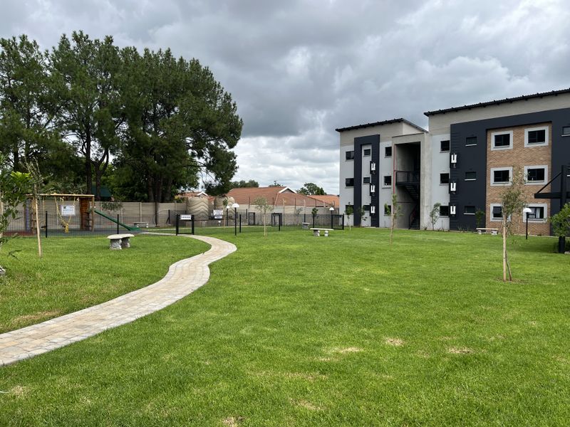 A well-maintained communal garden area featuring a paved walkway, manicured lawn, and perimeter fencing, situated within a modern townhouse complex.