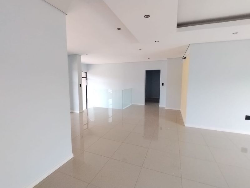 A spacious, modern open-plan living area featuring neutral porcelain floor tiles, recessed ceiling lighting, and a contemporary glass balustrade.
