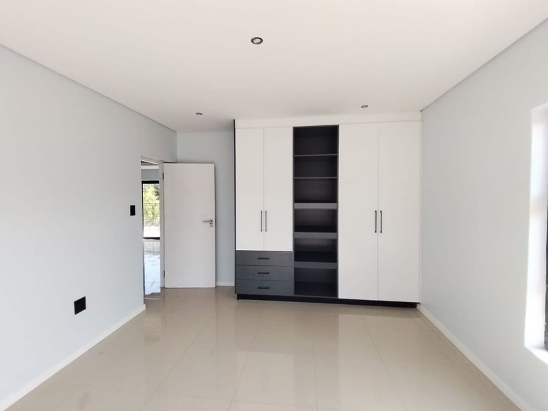 A bright, modern bedroom featuring neutral tiled flooring, recessed downlighting, and contemporary built-in cupboards with a mix of white and dark grey finishes.