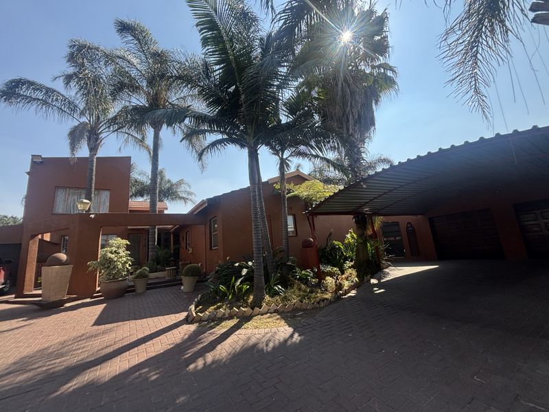 Front elevation of the house featuring a paved driveway, covered carport, integrated garage, and established garden with mature palm trees.