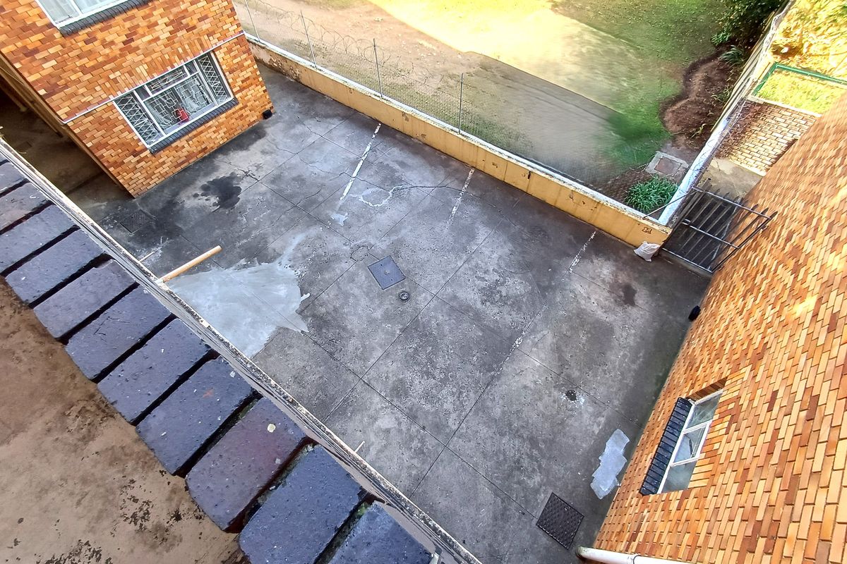 An elevated view of a paved rear courtyard area featuring brick building facades, security fencing, and basic drainage infrastructure.