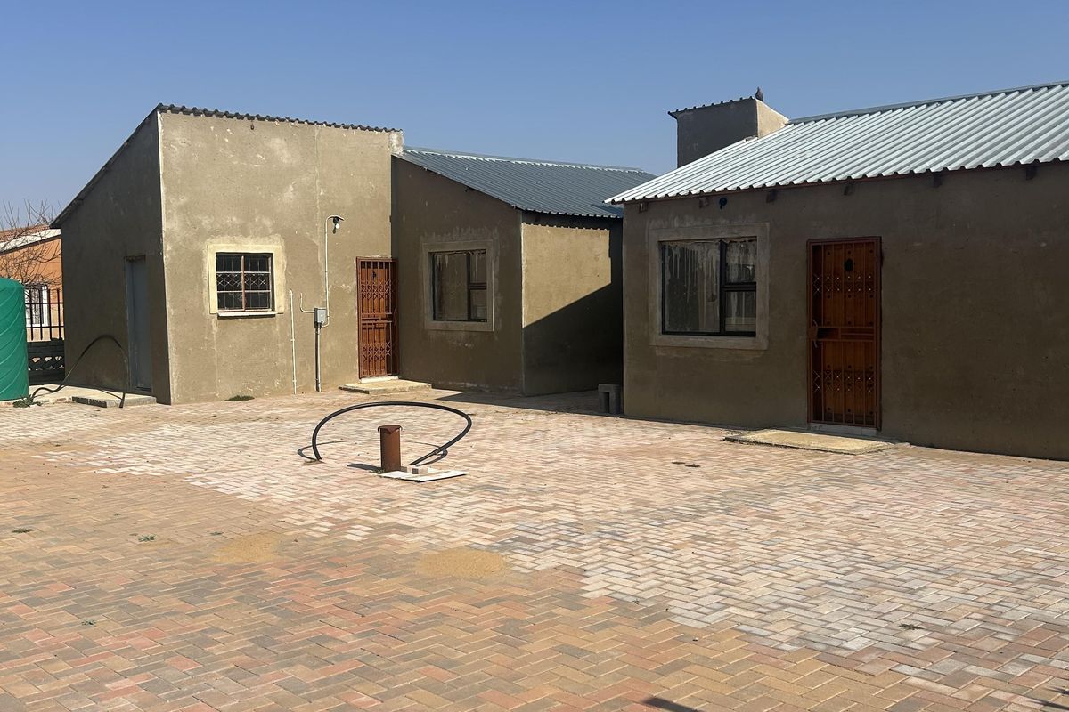 Rear view of the property featuring multiple structures with corrugated metal roofing, security gates, and a large paved courtyard area.