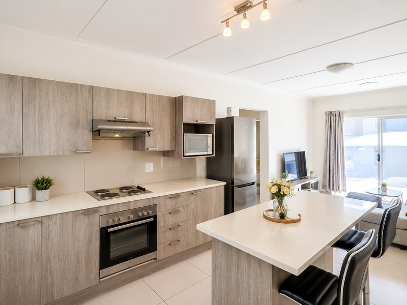 Modern open-plan kitchen featuring light wood-grain cabinetry, stone countertops, integrated appliances, and a central island, flowing into a bright living area with balcony access.