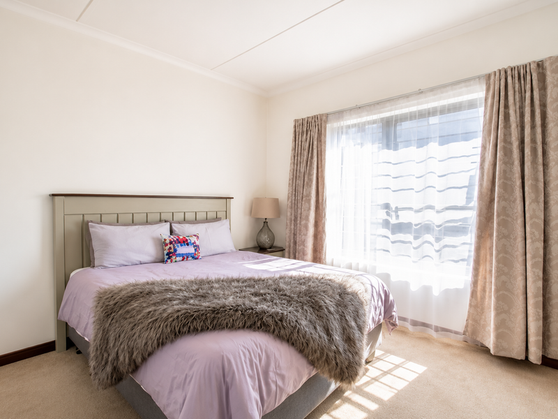 A well-maintained bedroom featuring neutral wall tones, carpeted flooring, and large windows with curtains allowing for natural light.