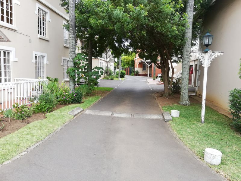A view of the apartment complex driveway featuring landscaped garden beds, mature trees, and paved access paths between the residential buildings.
