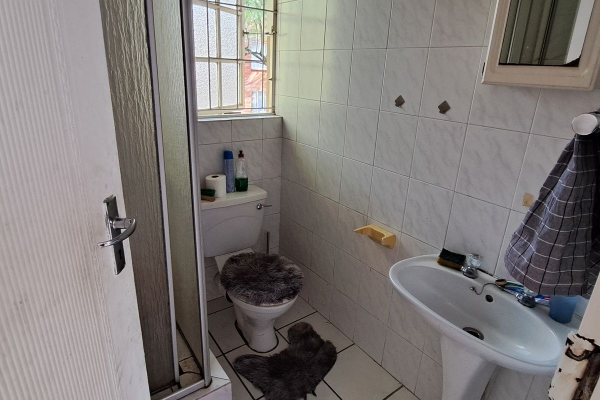 A compact bathroom featuring a shower enclosure, pedestal basin, and toilet, with tiled finishes and a window with security bars.