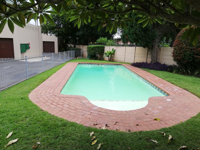 A communal swimming pool area featuring brick paving, landscaped lawn, and perimeter fencing within a residential complex setting.