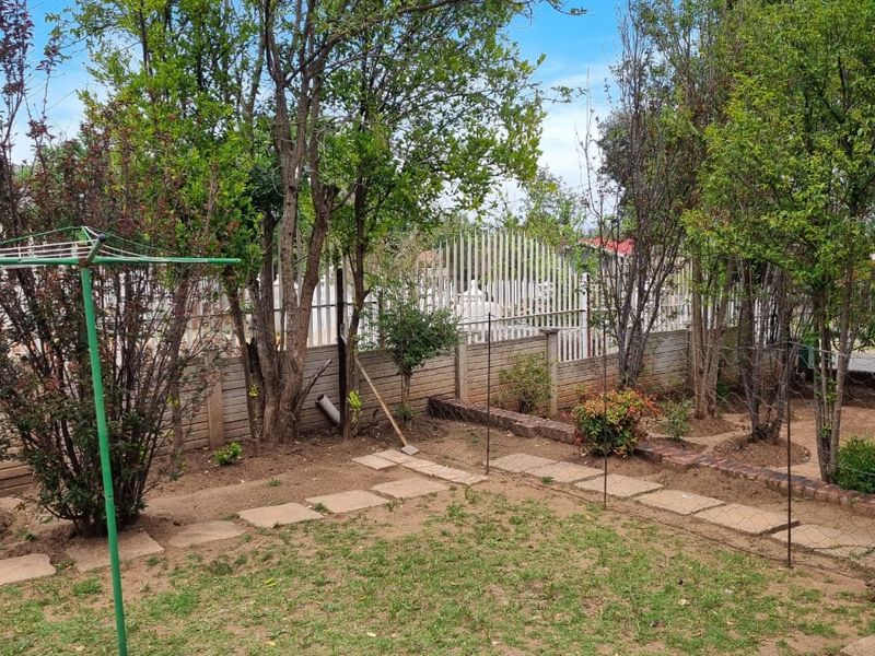 A private rear garden featuring mature trees, stone paving pathways, and perimeter security fencing with a precast concrete wall.