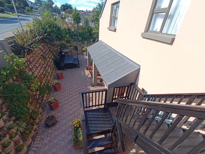 Rear exterior view featuring a paved patio area, covered walkway, and timber staircase leading to the upper level, set against a landscaped retaining wall.
