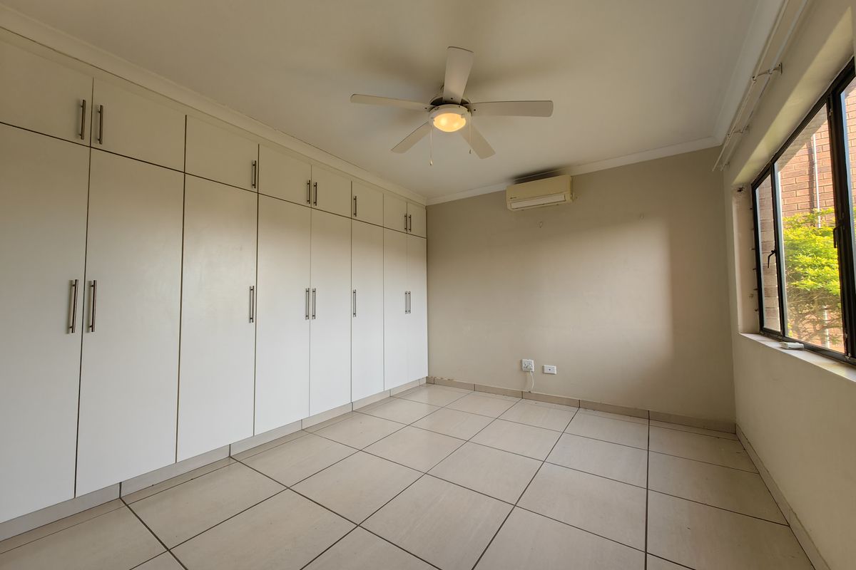 A bright, empty bedroom featuring extensive built-in cabinetry, a ceiling fan, and a wall-mounted air conditioning unit with neutral tiled flooring.