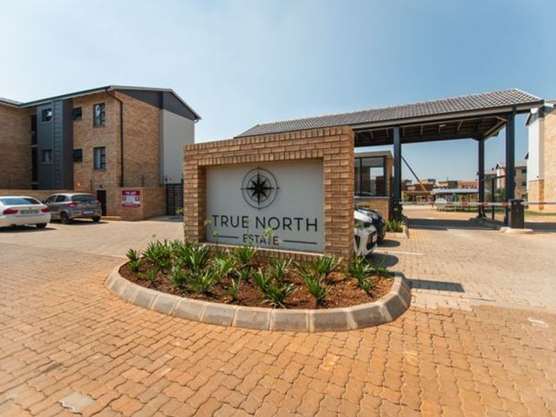 The entrance to True North Estate featuring a brick-paved driveway, security boom gate, guard house, and a prominent estate signage wall.