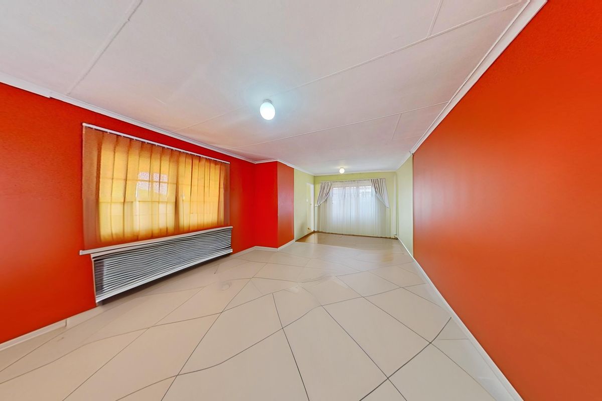 A spacious, empty living area featuring tiled flooring, bright orange accent walls, and large windows with fitted blinds.