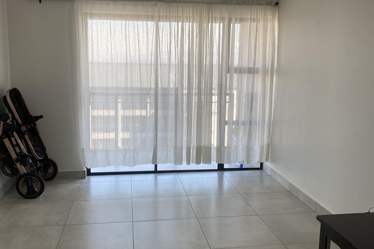 A bright, open-plan living area featuring neutral floor tiles, a large sliding glass door with curtain track, and access to an outdoor balcony.