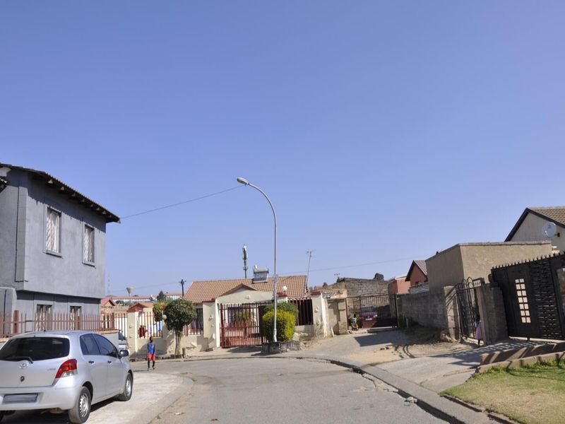 A street view showing the residential neighborhood approach with paved roads, boundary walls, and gated entrances to properties.