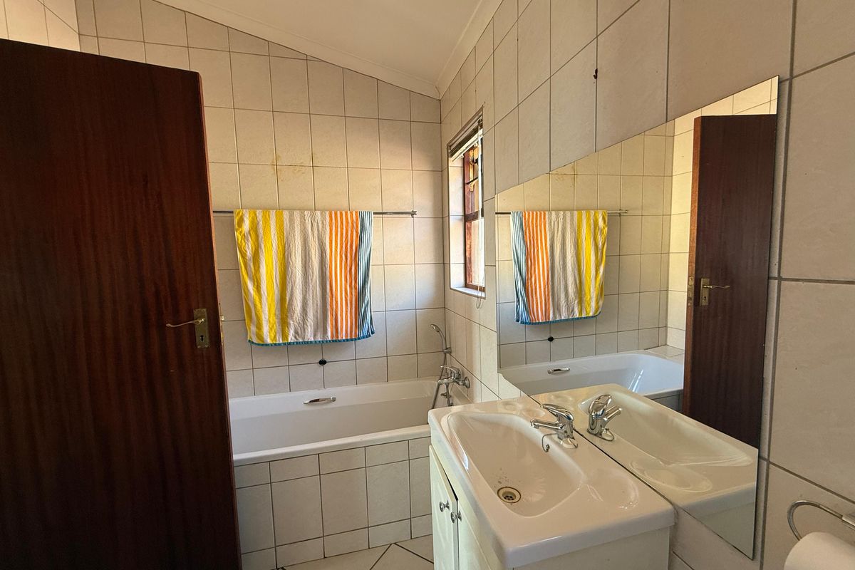 A functional bathroom featuring a built-in bathtub, vanity cabinet with basin, and wall-to-wall mirror, finished with neutral wall and floor tiling.