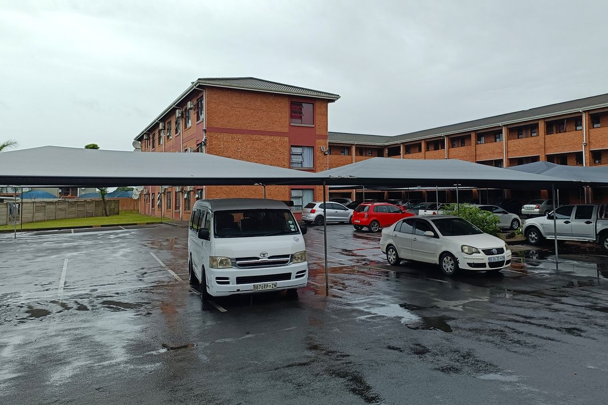 Exterior view of an apartment complex featuring a paved parking area with covered carports and brick building facades.