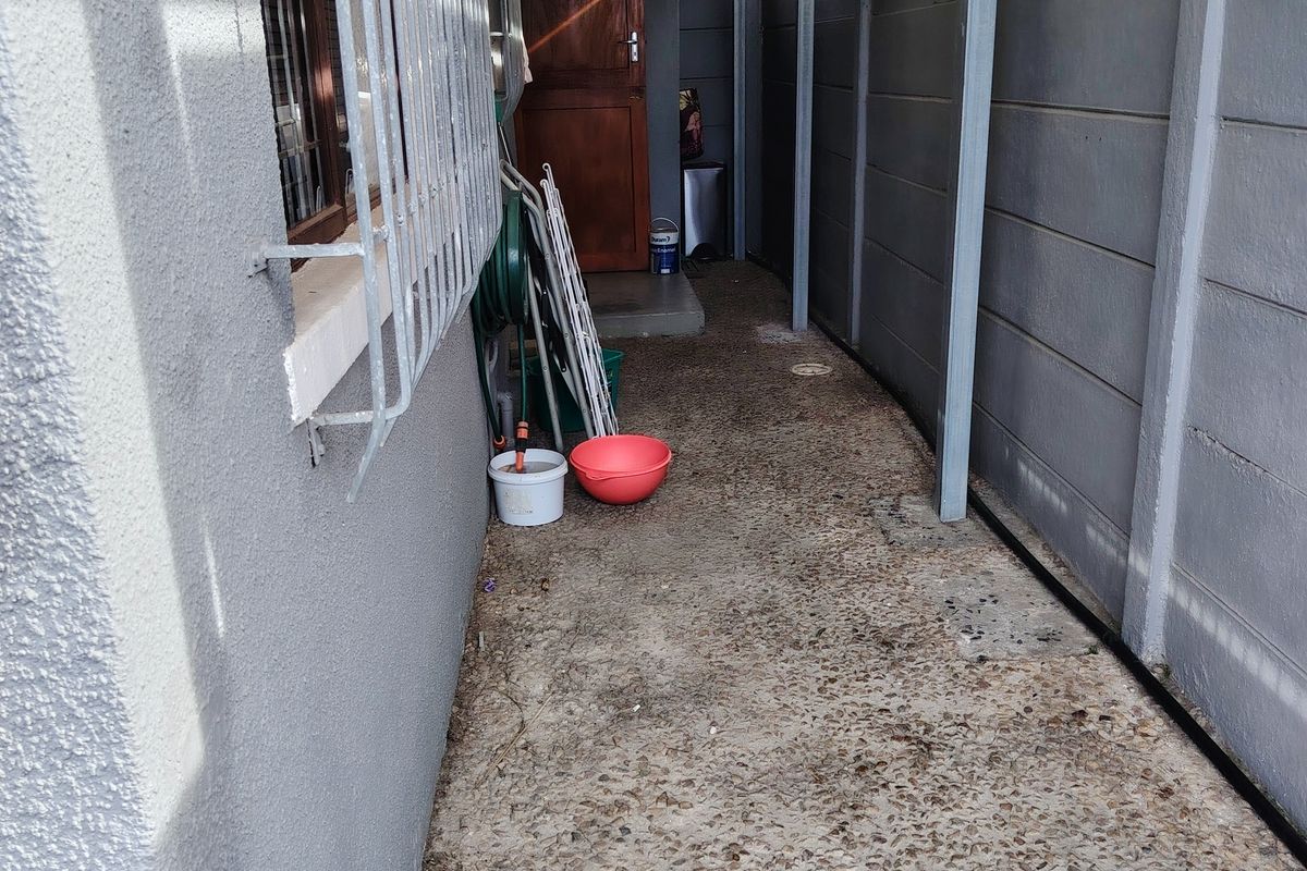 A narrow side passage featuring concrete paving, a precast boundary wall, and a covered walkway leading to a timber door.