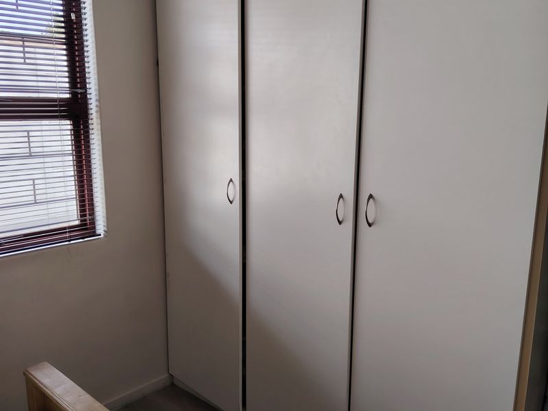 A bedroom featuring built-in white cupboards, laminate flooring, and a window fitted with venetian blinds.