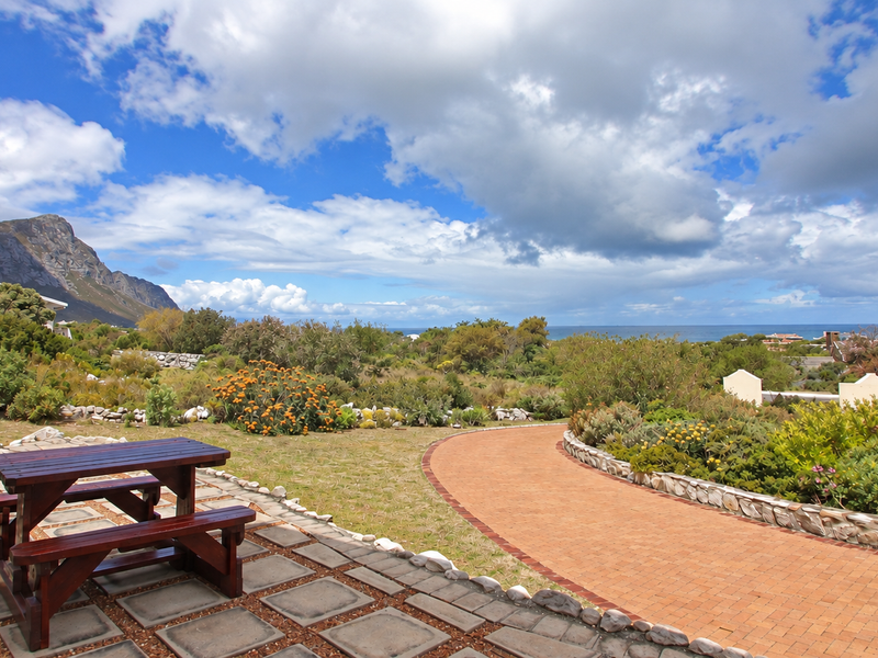 A spacious outdoor patio area featuring paved flooring and a brick pathway, overlooking a landscaped garden with scenic mountain and ocean views.