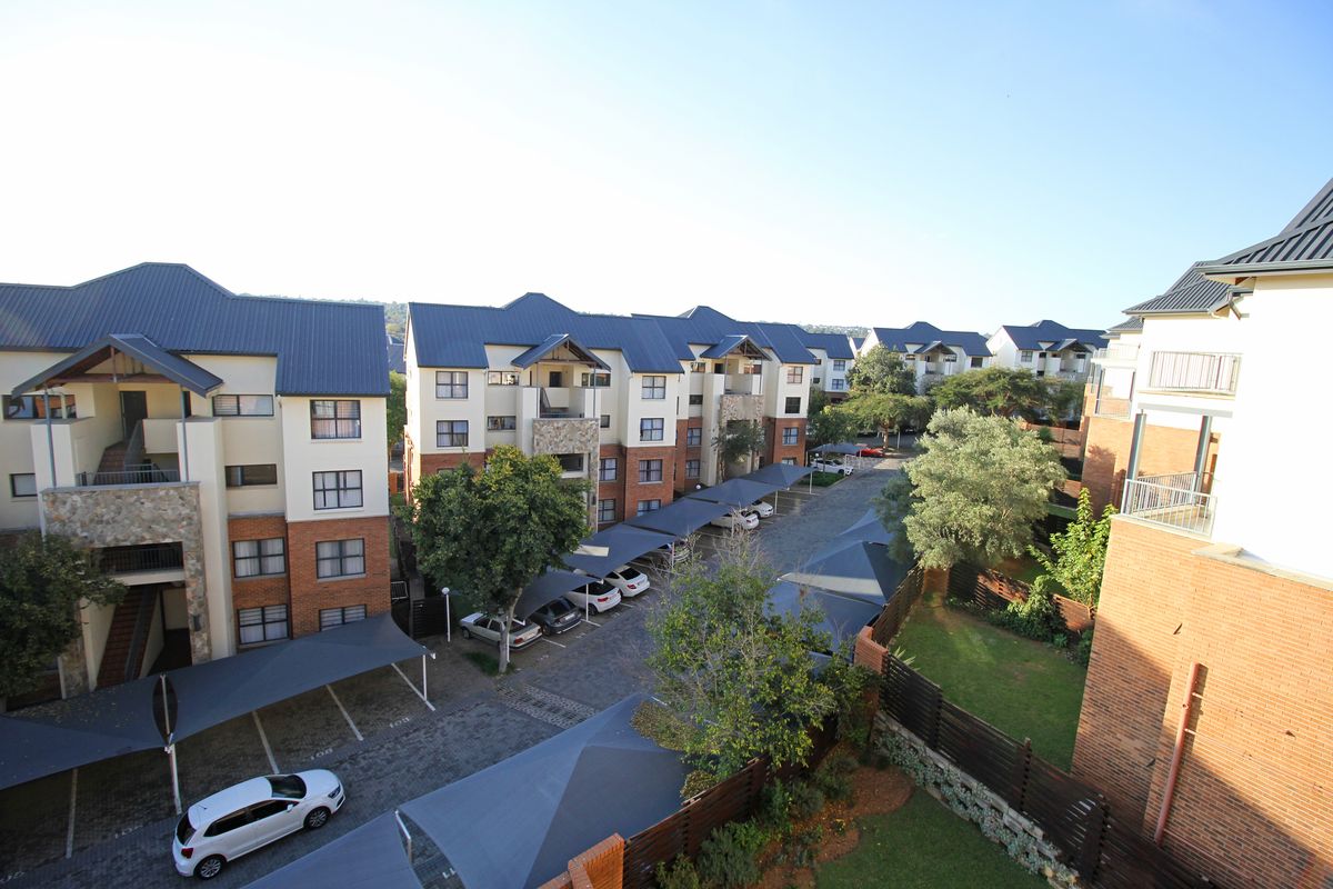 An elevated view of the apartment complex grounds, featuring paved parking areas with shade-cloth carports and landscaped garden spaces between the residential buildings.