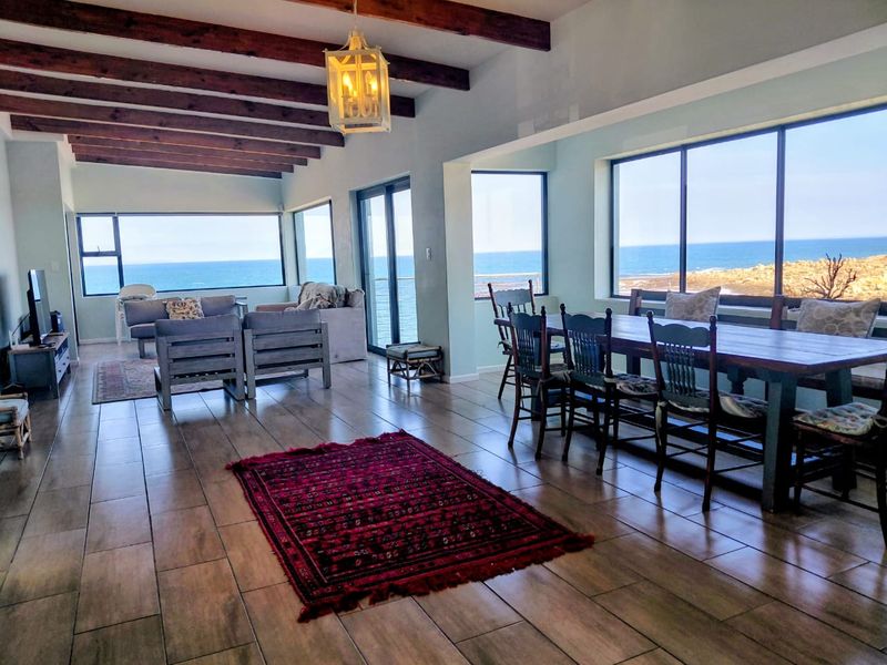 Spacious open-plan living and dining area featuring exposed timber ceiling beams, large format floor tiles, and expansive windows offering panoramic ocean views.